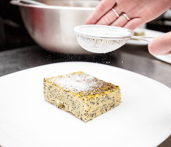 Image of a cake with poppy seeds in it being sprinkled with icing sugar through a sieve 
