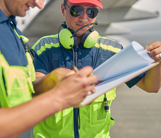 Image of 2 people in hivis work wear reading paperwork with an aeroplane in the background