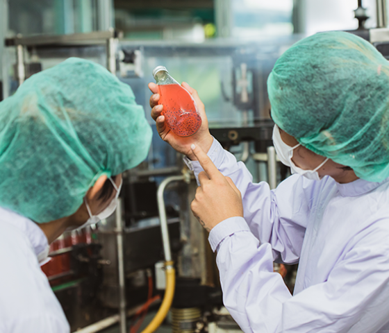 Image of people in a factory wearing hair nets and laboratory coats, looking at a salmon coloured drink in a glass bottle