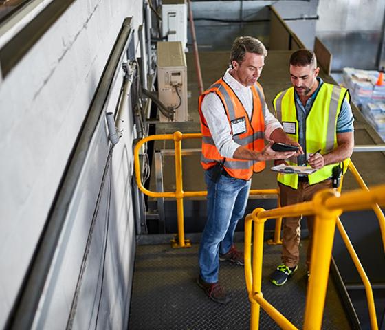 Image of 2 people in a warehouse looking at a clipboard and electronic device 