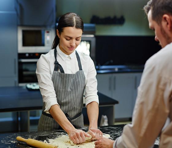 Image of 2 people in a commercial kitchen. one is kneading a dough, the other person is watching. 