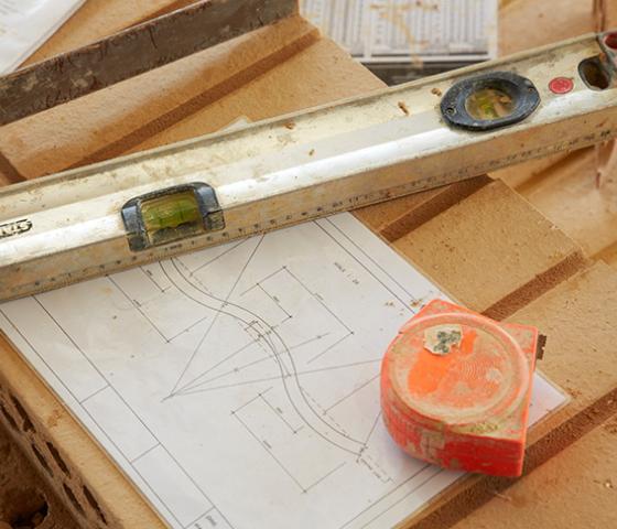 Image of building plans with a tape measure and level sitting on top of a pile of bricks 