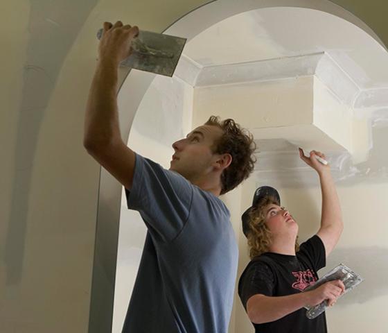 Image of 2 people using trowels to put white plaster on an unfinished wall 