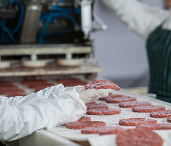 Image of meat burger patties on a conveyer belt being checked by a person wearing gloves 