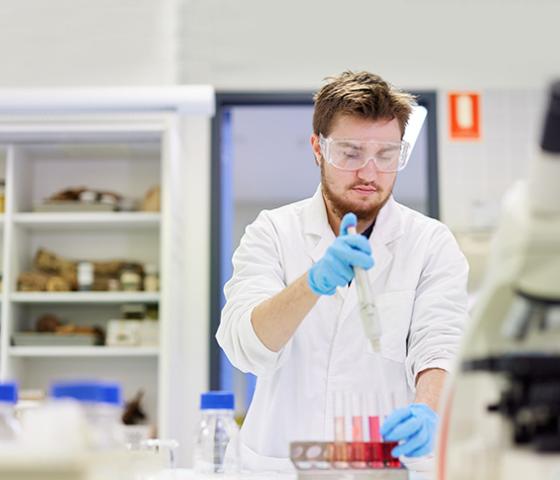 Image of a person wearing safety glasses and a lab coat. Person is using a baster and test tubes 